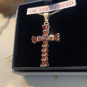 Op de afbeelding: Een met goud gevulde kruis hanger met rode edelsteen accenten. De hanger zit in een zwarte doos met een wit label waarop "18K Gold Filled" staat.