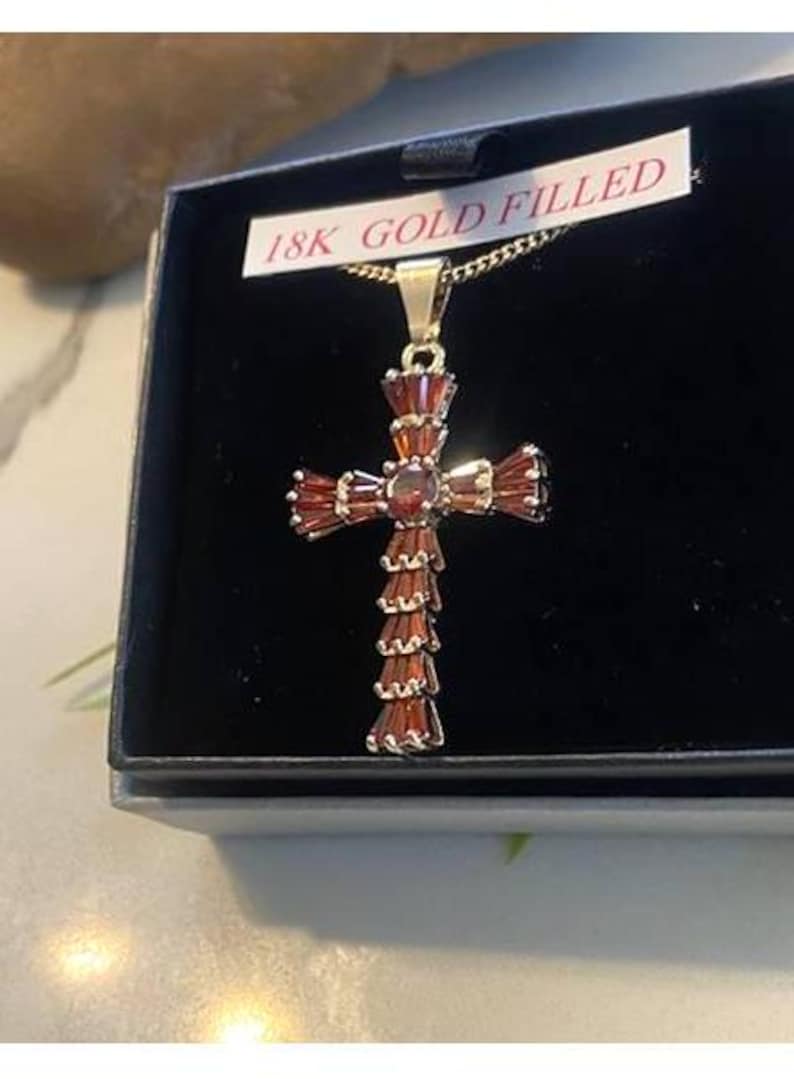 Op de afbeelding: Een met goud gevulde kruis hanger met rode edelsteen accenten. De hanger zit in een zwarte doos met een wit label waarop "18K Gold Filled" staat.