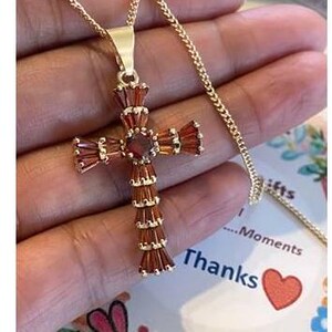 Op de afbeelding: Een gouden ketting met een rode edelsteen kruis hanger. Het kruis is gemaakt van kleine, rechthoekige stenen die in goud zijn gezet.