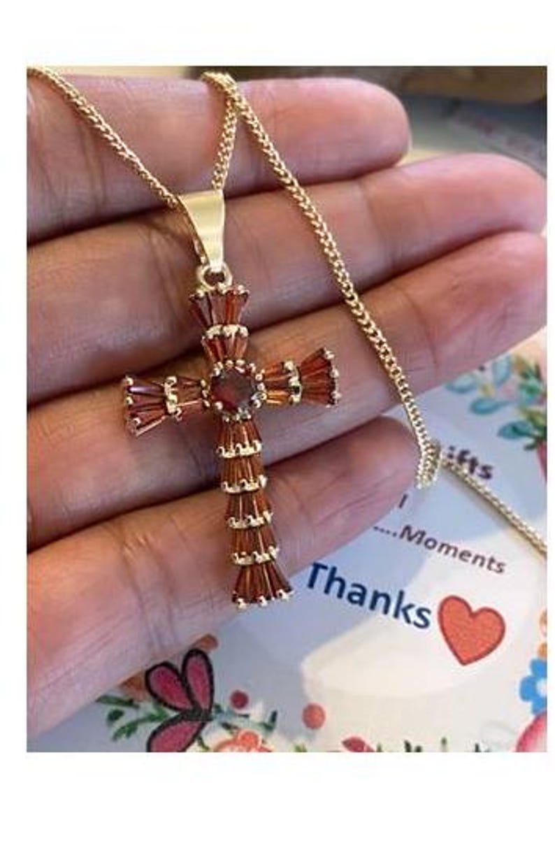Op de afbeelding: Een gouden ketting met een rode edelsteen kruis hanger. Het kruis is gemaakt van kleine, rechthoekige stenen die in goud zijn gezet.