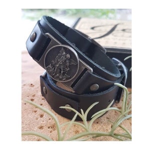 May include: Black leather bracelet with a silver metal charm featuring a religious figure. The bracelet has a buckle closure and is adorned with silver studs.