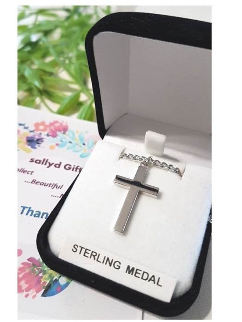Puede incluir: Un collar con una cruz de plata en una caja de terciopelo negro con una etiqueta blanca que dice "STERLING MEDAL".