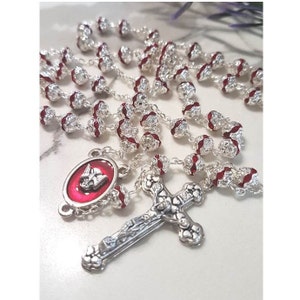 Può includere: Un rosario d'argento con dettagli rossi e un ciondolo a croce dettagliato. Le perline sono argentate con dettagli rossi. È presente anche un ciondolo ovale con sfondo rosso e un simbolo religioso.