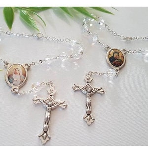 Op de afbeelding: Twee zilveren rozenkransen met heldere kristallen kralen en zilveren kruisbeelden. Elke rozenkrans heeft een zilveren ovale hanger met een religieus beeld.