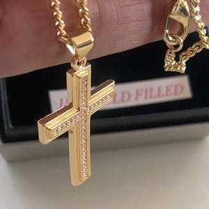 Op de afbeelding: Een gouden ketting met een gouden kruis hanger. Het kruis is versierd met kleine, heldere stenen. De ketting is een touwketting. De hanger is gemarkeerd met "Gold Filled".