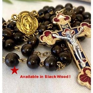 May include: A rosary with black wooden beads and gold-colored metal accents. The cross features a silver Jesus figure and red, blue, and gold details. The text "Available in Black Wood!" is visible.