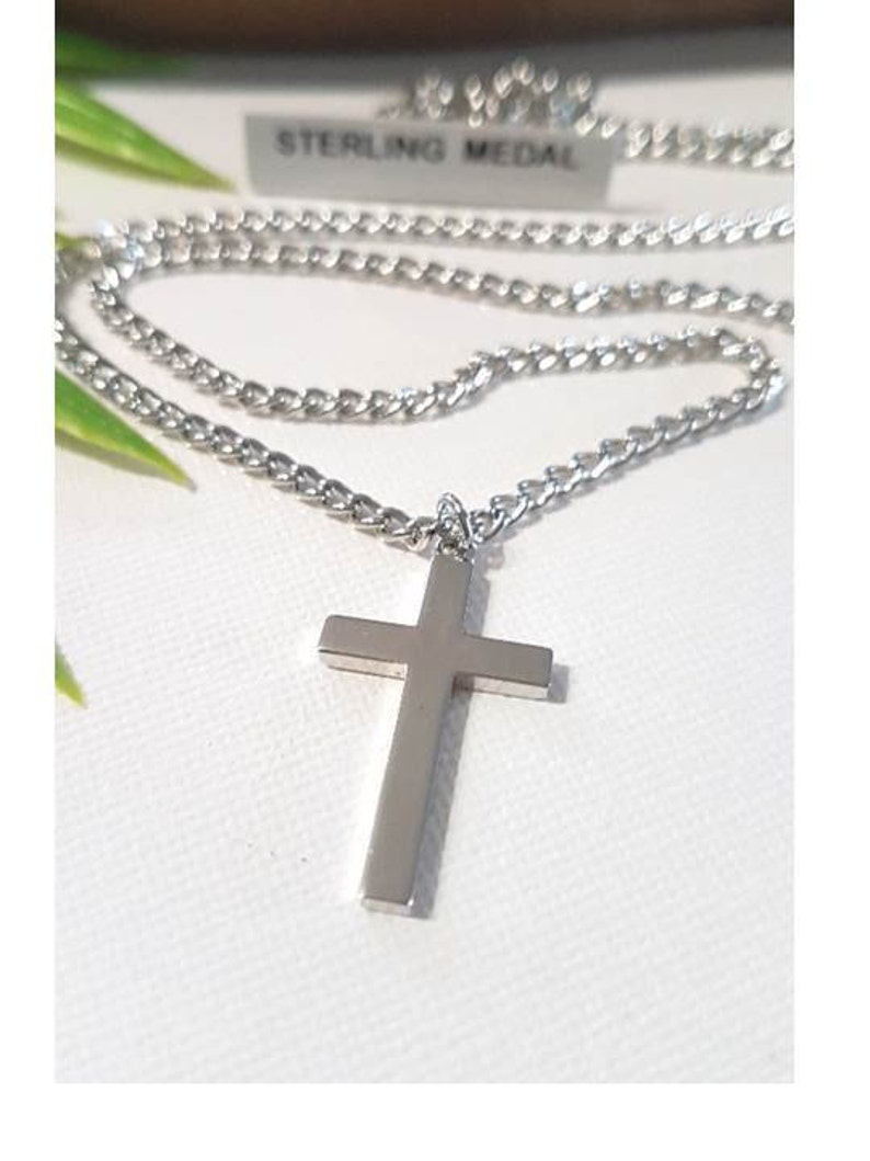 Puede incluir: Un collar de cadena de plata con un colgante de cruz de plata. La cadena es de estilo curb chain. La etiqueta en la cadena dice "Sterling Medal".