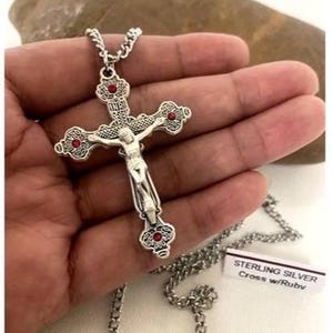 May include: A silver-colored cross pendant with a detailed crucifix figure and red accents. The cross is suspended from a matching chain. The pendant has the text "INRI" at the top. The tag reads "STERLING SILVER Cross w/Rubv".