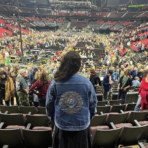 May include: A person wearing a blue denim jacket with a colorful, circular design on the back, standing in an arena filled with people. The jacket's design features a skull and lightning bolt motif. The arena has rows of seats and a stage.