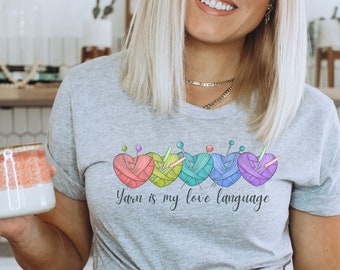May include: A woman wearing a gray t-shirt with a graphic of yarn balls and the text "Yarn is my love language". She is holding a mug in her left hand.