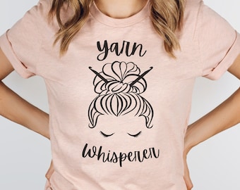 May include: A pink t-shirt with a black graphic of a woman's hair in a bun with knitting needles and the text "yarn whisperer".