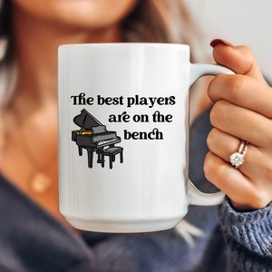 May include: White ceramic coffee mug with a black and white illustration of a piano and the text "The best players are on the bench".