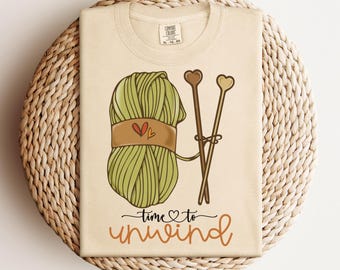 May include: Ivory t-shirt with a green ball of yarn, two knitting needles, and the text "time to unwind" in orange.