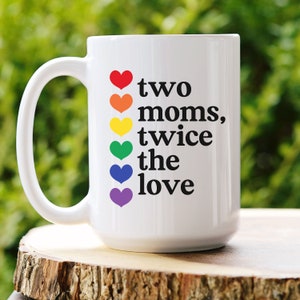 May include: White ceramic mug with a large handle. The mug features the text "two moms, twice the love" in black, with a rainbow of hearts. The mug is sitting on a wooden surface.