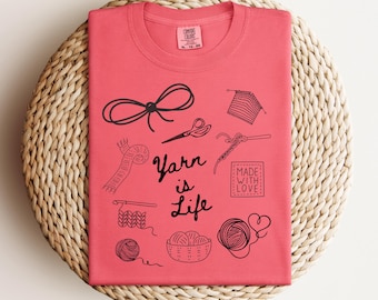 May include: A pink t-shirt with a black graphic design that says "Yarn is Life". The graphic includes various yarn-related items, such as balls of yarn, knitting needles, and scissors.