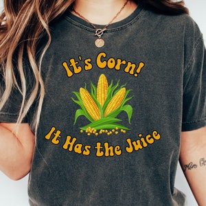 May include: A dark gray t-shirt with the text "It's Corn! It Has the Juice" in yellow letters. A cartoon image of corn is in the center of the shirt.