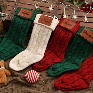 Peut inclure: Plusieurs bas de Noël personnalisés, verts, rouges et crème. Chaque bas porte une étiquette en cuir avec un nom. Un ours en peluche et une boule rayée rouge et blanche complètent la scène festive.