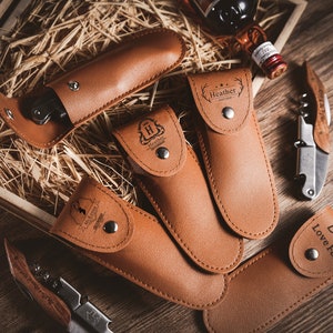 Puede incluir: Un conjunto de cinco estuches de cuero marrón con grabados personalizados. Cada estuche contiene una herramienta diferente, incluyendo una navaja de bolsillo, un sacacorchos y un abridor de botellas. Los estuches están dispuestos sobre una superficie de madera con relleno de paja.