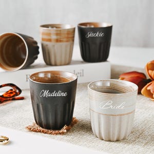 May include: A collection of ceramic cups with personalized names, including "Jackie," "Madeline," and "Bride." The cups have a ribbed design and a two-tone color scheme of black, beige, and brown. The cups are arranged on a neutral surface with decorative elements.