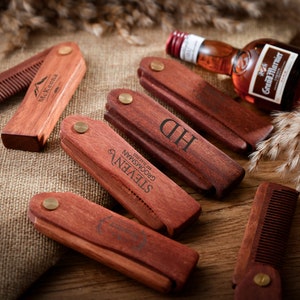 May include: A set of six wooden pocket combs with personalised engravings. The combs are made of dark red wood and have a brass hinge. The engravings include names and the words "Groom" and "Groomsman".