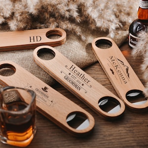 Könnte beinhalten: Vier Flaschenöffner aus Holz mit personalisierten Gravuren. Die Öffner haben an einem Ende einen Flaschenöffner und am anderen Ende einen Flaschenverschluss-Halter. Die Gravuren beinhalten Namen, Daten und das Wort "Groomsmans".