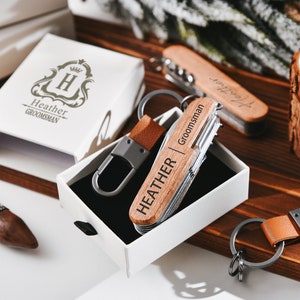 May include: A personalized wooden pocket knife with a leather keychain. The knife has the name "Heather" and the word "Groomman" engraved on it. The knife is in a white box with a black interior.