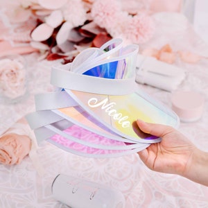 May include: Stack of iridescent visor hats with white trim and the name "Nicole" written on the front.