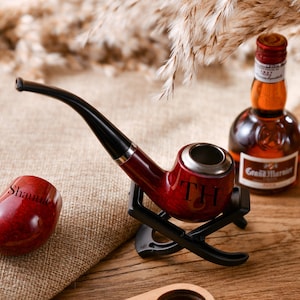 Könnte beinhalten: Eine rote Holzpfeife mit schwarzem Schaft und silberner Schale. Die Pfeife ist mit den Initialen "TH" graviert und steht auf einem schwarzen Ständer. Eine kleine Flasche Grand Marnier Likör ist ebenfalls im Bild zu sehen.