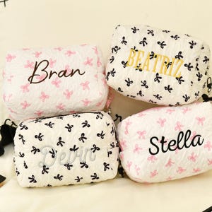 May include: Four cosmetic bags with personalized names. The bags are white with a textured pattern and decorated with pink or black bow accents. The names "Bran", "Beatriz", "Devin", and "Stella" are embroidered on the bags in various colours.