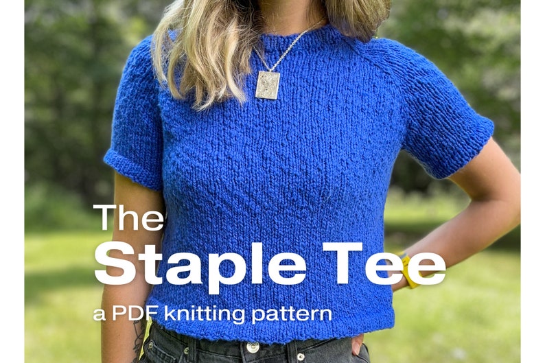 A relaxed fit knitted tee knitting in worsted weight blue yarn worn on a female model who is standing outside. TEXT: The Staple Tee, a PDF knitting pattern