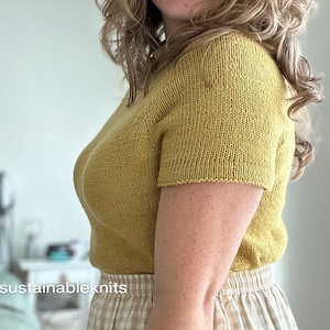 A relaxed fit knitted tee knitting in dull yellow  DK weight yarn worn on a female model and tucked into a gingham checked skirt.