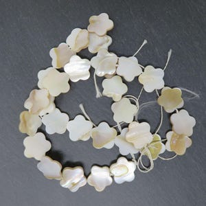 May include: Strands of flower-shaped mother-of-pearl beads. The beads are a creamy white color with iridescent highlights. The beads are strung on white string, and are suitable for jewelry making.