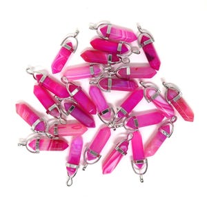 May include: A collection of pink and magenta crystal pendants with silver-colored metal caps and loops. The crystals have a polished, elongated shape with visible banding. The pendants are scattered on a white background.