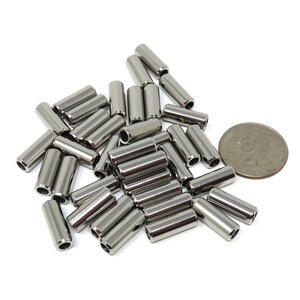 May include: A collection of shiny, cylindrical metal beads, scattered on a white surface. The beads are silver in color and have a smooth, polished finish. A U.S. quarter is included for size comparison. These beads are likely used for jewelry making or crafting.