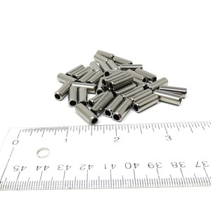 May include: A pile of small, cylindrical metal tubes with a hollow center. The tubes are silver in color and appear to be made of stainless steel. A ruler is in the foreground, with measurements in inches.