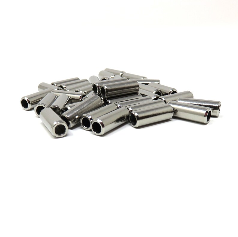 May include: A pile of shiny, silver-colored metal tubes with a cylindrical shape. Each tube has a hole at both ends. The objects are scattered on a white background, creating a clean and simple aesthetic.