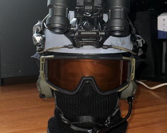 May include: A tactical helmet equipped with dual-tube night vision goggles mounted on the front, amber-tinted protective goggles, a black balaclava covering the lower face, and a communication headset with a boom microphone attached to the side.