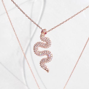 Może przedstawiać: Złoty naszyjnik z zawieszką w kształcie węża. Wąż jest pokryty małymi, przezroczystymi kamieniami i ma czerwone oczy.