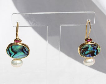 May include: A pair of gold earrings with a round abalone shell and a pearl drop. The earrings have a small pink stone accent.