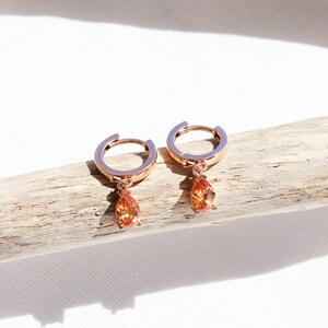 May include: A pair of rose gold hoop earrings with a teardrop-shaped orange gemstone dangling from each hoop.