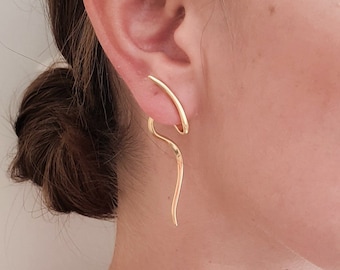 May include: A pair of gold-toned earrings with a unique, abstract design. The earrings are made of a thin, curved metal that creates a flowing, organic shape. The earrings are worn by a person with their eyes closed.