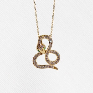 Może przedstawiać: Złoty naszyjnik z wisiorkiem w kształcie węża z delikatnym łańcuszkiem. Wisiorek w kształcie węża jest inkrustowany małymi, błyszczącymi kryształami i ma zielony kamień szlachetny w oku. Naszyjnik jest prezentowany na białym tle.