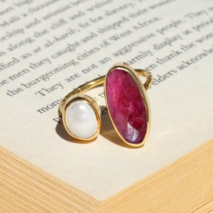 Puede incluir: Un anillo de oro con dos piedras: un gran rubí ovalado facetado y una perla blanca redonda más pequeña. El anillo está sobre un libro abierto con texto.