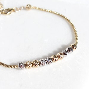 Puede incluir: Una delicada pulsera dorada con un dise&ntilde;o de cadena. La pulsera presenta una serie de cuentas esf&eacute;ricas texturizadas en tonos dorados y plateados. El cierre y la cadena son dorados, creando un sutil contraste sobre un fondo blanco.