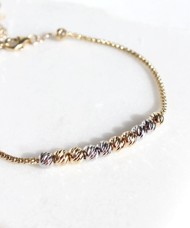 Puede incluir: Una delicada pulsera dorada con un dise&ntilde;o de cadena. La pulsera presenta una serie de cuentas esf&eacute;ricas texturizadas en tonos dorados y plateados. El cierre y la cadena son dorados, creando un sutil contraste sobre un fondo blanco.