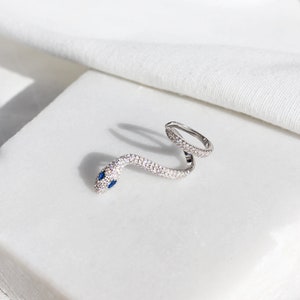 May include: A silver snake ring with a wrap-around design. The snake's body is encrusted with small, clear crystals, and its head features two small blue gemstones for eyes. The ring is displayed on a white surface.