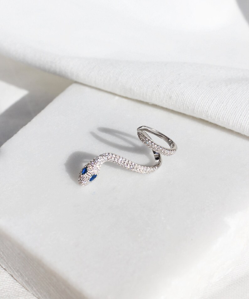 May include: A silver snake ring with a wrap-around design. The snake's body is encrusted with small, clear crystals, and its head features two small blue gemstones for eyes. The ring is displayed on a white surface.