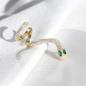 May include: A gold-colored snake ring with a coiled design. The ring is encrusted with small, clear crystals along its body. The snake's head features two small, green emerald eyes. The ring is displayed on a white surface.