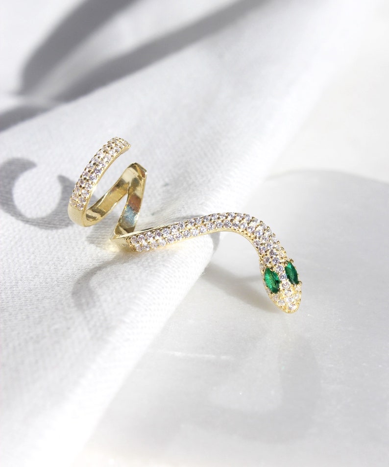 May include: A gold-colored snake ring with a coiled design. The ring is encrusted with small, clear crystals along its body. The snake's head features two small, green emerald eyes. The ring is displayed on a white surface.