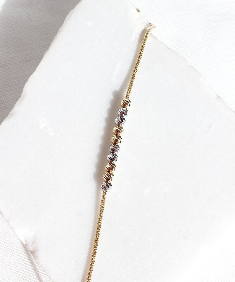 Puede incluir: Una delicada pulsera de cadena dorada con una serie de cuentas esf&eacute;ricas texturizadas en oro, plata y oro rosa. La pulsera se muestra sobre una superficie blanca, mostrando su intrincado dise&ntilde;o y variaciones de color. Esta joya es adecuada para el uso diario.